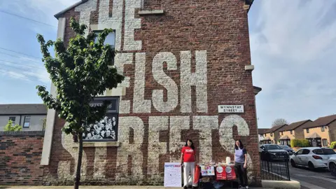 Acorn handout Two female campaigners against the Welsh Streets rent rises stand at a table with leaflets under a gable end terrace with the words 'The Welsh Streets' written on the wall. A tree is to their right in front of the house.