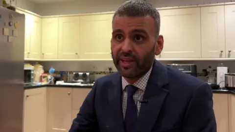 A man wearing a navy blue suit and tie, with a white shirt and blue lines sits at a table, looking towards a camera. A kitchen surface top and utensils can be seen behind him.