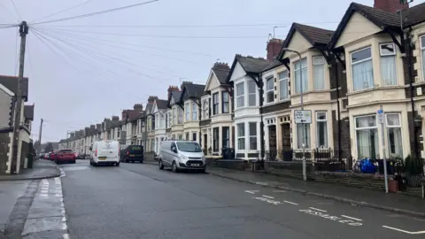 Houses line the street on both sides of Moorland Road in Splott, Cardiff. Cars and vans are dotted along the road on either side and a white van is seen driving along the road, away from the camera. A disabled parking bay with the words 'disabled' and 'anabl' can also be seen on the right.