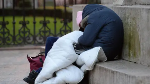 PA Media A person sleeping rough slumped against some stone steps. They're wearing a navy blue winter coat and have a white blanket covering their lap.