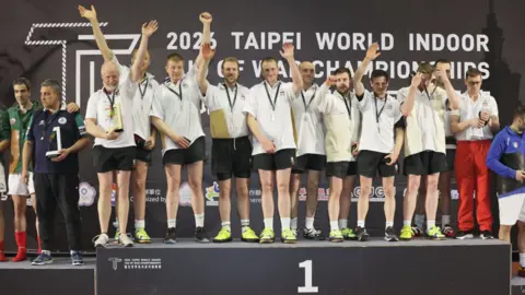 Christine Yang / Chinese Taipei Tug of War Association The team, dressed in white T'shirts and black shorts, stand on a large, black podium with their medals around their necks with some waving.