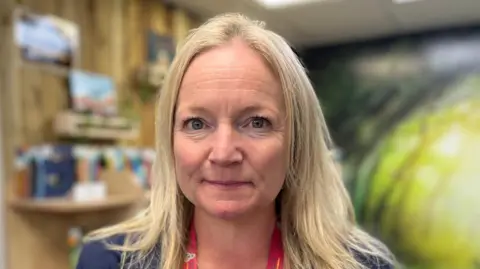 A lady with long blonde hair and grey/blue eyes smiling slightly and looking straight at the camera. She is wearing a nay blue jacket and a red lanyard. There is a school library blurred in the background.