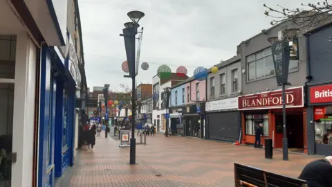 The pedestrianised street is bordered by various shop fronts, including a red shop with a sign that reads: "The Blandford". Red and grey bricks on the road are arranged in a zigzag pattern. Above, large, colourful circular bunting is hanging. There are people coming in and out of the shops.
