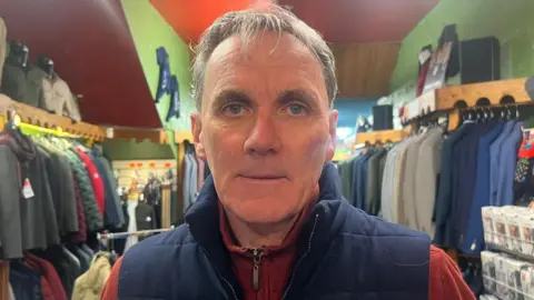 A man in a blue gilet and an orange fleece stands in the middle of men's clothes shop. A number of suits and items are on rails behind him.