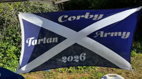 Emma Baugh/BBC A blue and white flag showing the Scottish saltire – a white diagonal cross on a blue background. Written on it is "Corby Tartan Army 2026" in white block capitals.