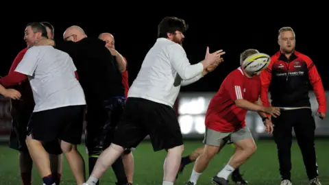 Krishan Turner-Dave Seven members of the club mid-game. The scrum half is passing the ball from the back of a touch rugby scrum or ruck.