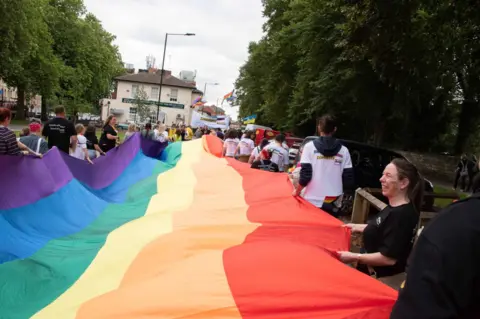 Doncaster Pride/Malcolm Johnston People holding a long rainbow flag stretched horizontally between them as they parade through a Doncaster street