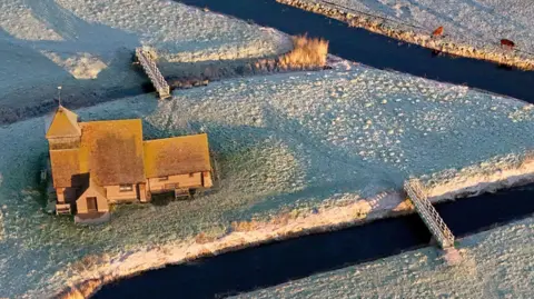 An aerial shot of a church which sits next to a winding river through a frosty field. There are deer at the banks and a bridge.