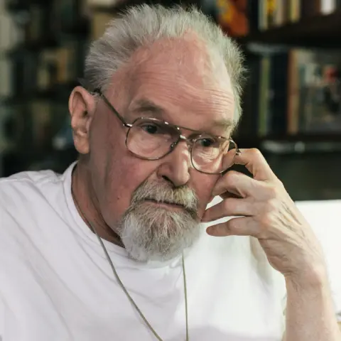 PA Media Alasdair Gray, a grey-haired man with a grey beard and moustache, stares solemnly to the side, his left hand at his cheek, wearing a plain white t-shirt and thin-rimmed glasses.