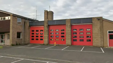 Hexham's former fire station. It is a two-storey brick building with an appendage to the right, with three large doors, for fire engines to enter and leave through. There is a car park to the front of it.
