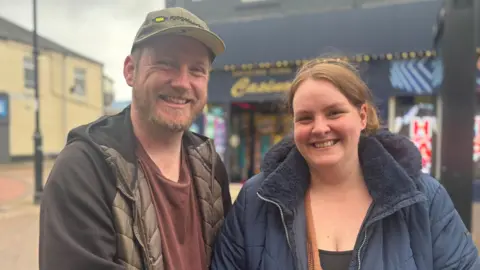 Ron and Hazel are standing in front of a shot on a high street in Widnes. They are both smiling at the camera. 