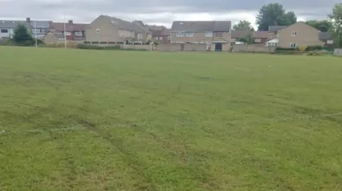 LDRS Rugby posts and several houses at a grassy sports pitch marked with tyre marks from illegal off-road bikes driving on sports pitches. 