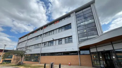 Ellen Knight/BBC Wide-angle photograph of the building that will be refurbished. It is four storeys high, clad in grey and with large glass windows. The sky is blue with grey clouds. 