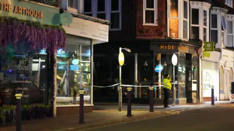 Eddie Mitchell A police officer stands in front of shops cordoned off by police tape. 