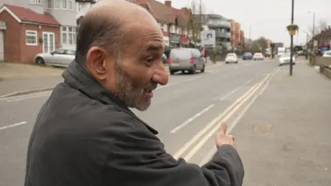 A man with brown skin and a grey stubbled beard standing on the pavement next to a main road in north London