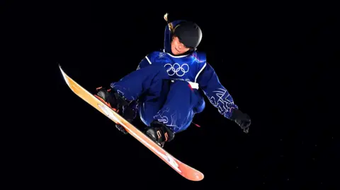 PA Media Maisie Hill during the Women's Snowboard Big Air Qualification. She is in the air with her snowboard tilted at a near 45 degree angle. Her knees are bent and one of her hands is holding the back edge of her board while the other is horizontal to her body. She is wearing blue snowboarding gear and the bottom of her board is orange. The background is black. 