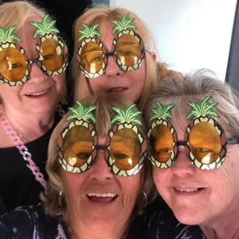 Susie Matthews Four smiling older women pose together for a selfie, each wearing oversized novelty sunglasses shaped like pineapples.
