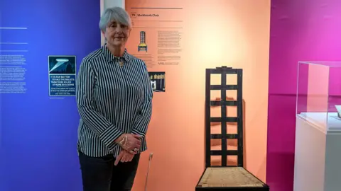 West Northamptonshire Council A woman standing in the exhibition with her hands crossed in front of her. She is wearing dark trousers, a striped top and short grey hair. She is standing by a black and wicker chair. 