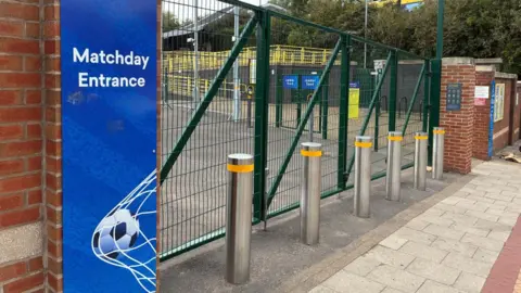 A blue sign with a football hitting a net saying "Matchday entrance" next to a set of grey metal bollards and a closed gate. Through the wire gate you can see a rail surrounding a walkway leading to a railway station platform.  