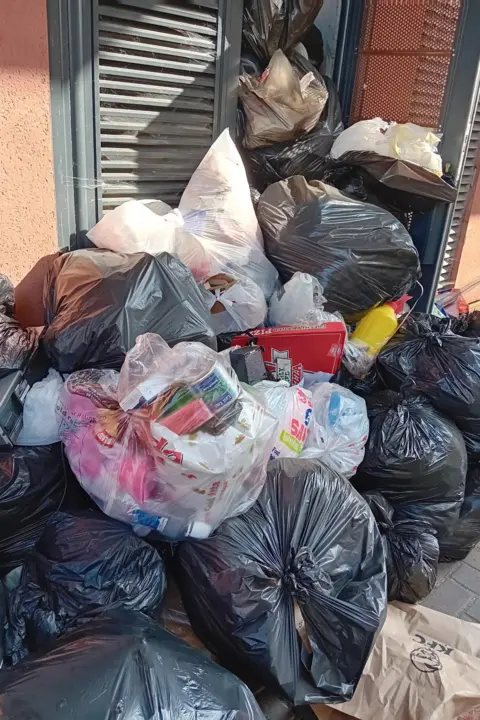 LDRS Large accumulation of tightly packed bin bags and mixed rubbish piled high against a building wall, with cartons and loose items visible, obstructing the walkway.