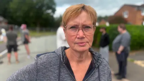 A woman with short ginger hair, large glasses and a grey top in a street with the blurred shapes of people behind her 