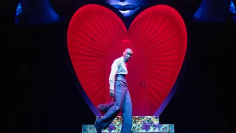 Daniel Boud Cynthia Erivo stands in front of a heart on stage in a suit while a visual projection of her face is above it. 