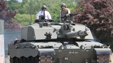 PA Media King Charles has the top half of his body sticking out of the top of a tank, he is wearing a white shirt with a red and green tie and some sunglasses. He is also wearing a green helmet with ear defenders and a microphone. Beside him is a man in camouflaged military uniform who is holding a gun and wearing the same style of helmet, ear defenders and microphone as the King.