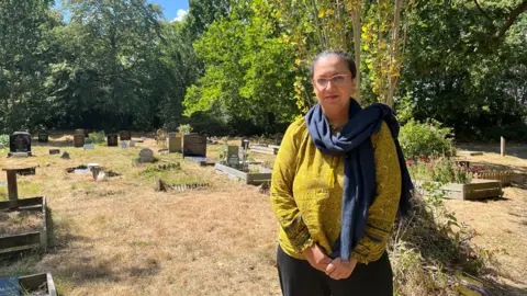 Kumail Jaffer/LDRS A lady with long hair tied back and glasses, wearing a yellow/green jumper and blue scarf poses for a photo in a graveyard on a sunny summer's day.