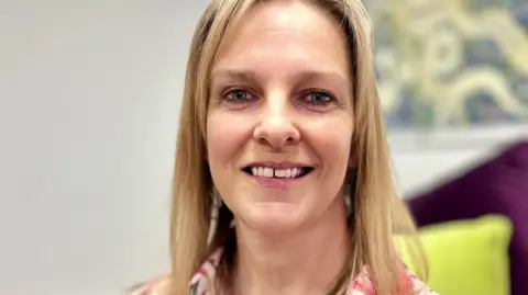 Portrait photograph of Becky Ward smiling to camera. She has long blonde straight hair and is wearing a floral blouse.