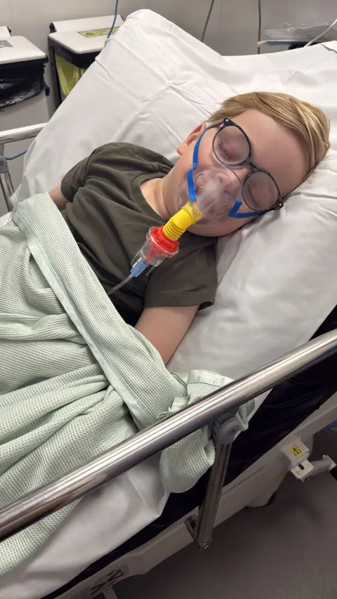 Supplied Finley in a hospital bed wearing a green t-shirt. He is wearing glasses and has a mask on his face. 