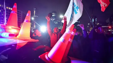 Getty Images A man holds his hands in the air, in front of crowds of people waving flags, while in the foreground a police car covered in orange hazard cones flashes red and blue lights