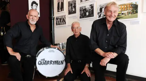 Getty Images Len Garry, Rod Davis and Colin Hanton are all pictured wearing black shirts and black trousers. They are crouched next to a drumkit with "The Quarrymen" written in black writing on a white bass drum skin