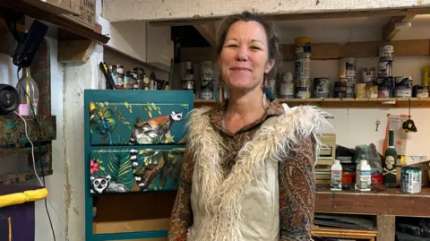 Nienke has dark hair and wears a patterned top and fluffy trimmed body warmer. She is standing in her workshop next to a chest of drawers being transformed with a jungle style motif. 