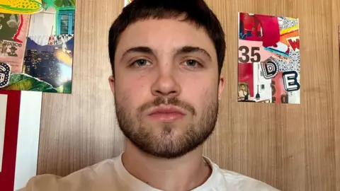 Christian Lotz has a dark brown beard and hair and is wearing a white T-shirt. He is looking directly at the camera. There are six brightly coloured red, and green portrait shaped collage-style artworks on the wall behind him, which has a brown timber-effect.
