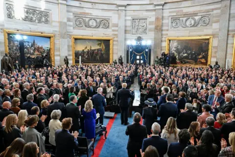 Getty Images Trump stands at a podium in the Rotunda which is full of guests. He has his back to us and appears to be making a celebratory gesture with his right fist aloft. 