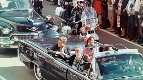 Getty Images President Kennedy, pictured riding in an open convertible minutes before he was shot by a sniper. He is accompanied by his wife, who is wearing pink, and police officers. The are crowds of people lining the streets to see him