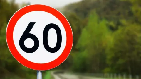 A circular 60mph speed limit sign with a black 60 in a circle with a red ring around it. A blurred curvy round can be seen behind it surrounded by trees.