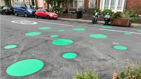 A cluster of large, green spots can be seen painted on a road. A 20 mph speed limit is also painted on the surface. A row of houses can be seen - and several vehicles parked at the kerbside. 