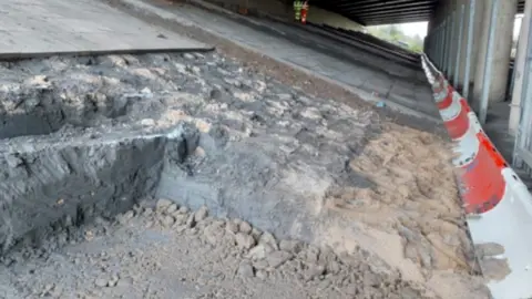 National Highways This picture was taken underneath the M5 bridge. The concrete underneath the bridge appears worn away. There is a red and white low barricade in front of the damaged concrete. 