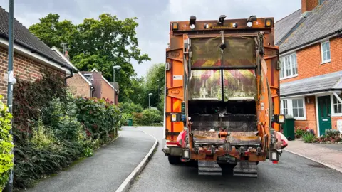 The rear of an orange bin lorry working in a cul-de-sac. Red-brick houses are on either side.