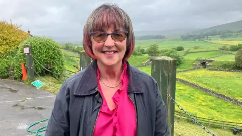 A woman, Jill, wears a pink top and grey jacket. Her hair has pink highlights. She stands in a car park, overlooking a picturesque landscape of fields and trees, looking down a valley.