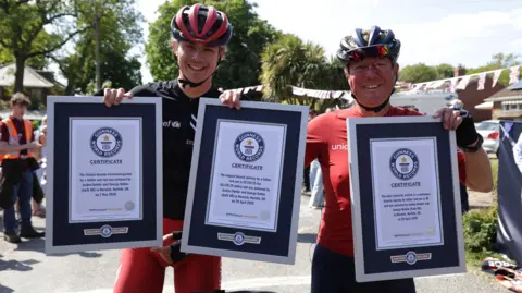 Josh and George Kohler are standing next to each other. They are both wearing red and black cycling wear and helmets. They are holding up three Guinness World Record certificates.