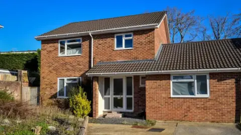 A detached red brick house with driveway and white PVC windows. Under a blue sky.