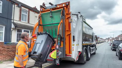 A garbage collector, wearing a bright orange hi-vis vest and red gloves,  emptying a black bin into the back of a large orange and white waste collection truck on a residential street.