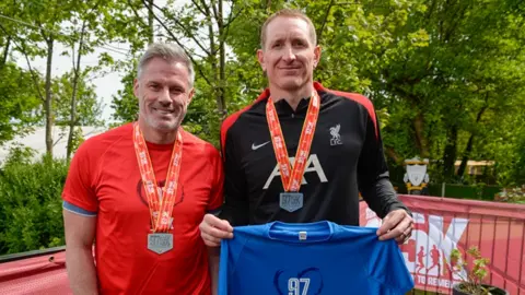 Former Liverpool stars Jamie Carragher and Chris Kirkland wearing running gear and Run for the 97 medals in Liverpool's Stanley Park. Kirkland is holding up a blue Run for the 97 shirt. They are both smiling.