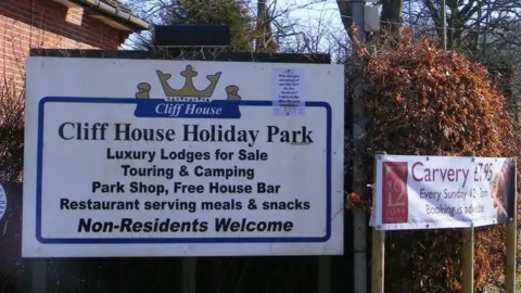 A sign at the entrance of Cliff House Holiday Park. It is white with lettering in black font advertising various aspects of the park, including a bar, shop and restaurant.