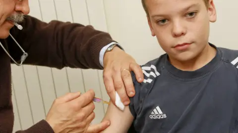 A child with a grey t-shirt and a pair of hands putting a needle into his arm