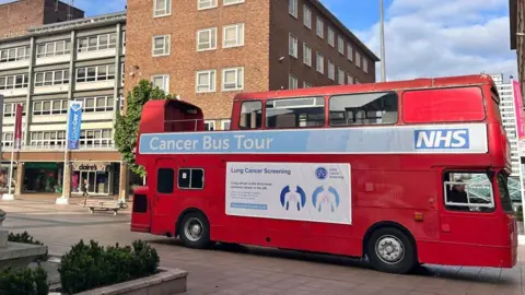 A red open-top London bus that has been converted into a cancer screening information service. It is parked up in a city centre in front of shops. It has a big sign that reads "Cancer Bus Tour". 