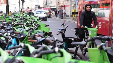 A person on a Lime bike in central London, with a red bus in the background, and a densely parked row of e-bikes in the foreground. 