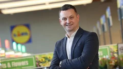 Lidl Northern Ireland Shows a man in a blue jack, white button-down shirt and arms folded standing in a shop with Lidl sign in the background.